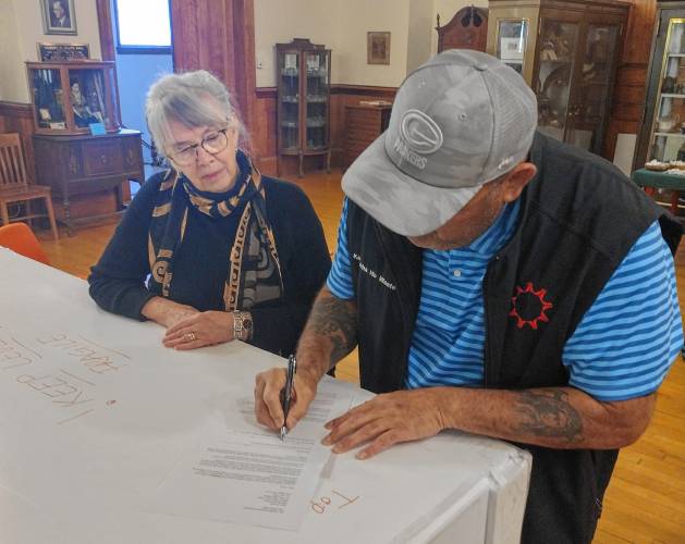Barre Museum Association board member Elizabeth Martin looks on as Fifth Member and Tribal Historic Preservation Officer of the Oglala Sioux Tribe Justin Pourier signs the agreement between the Woods Memorial Library and the tribe regarding the return of items to the Pine Ridge Reservation in South Dakota. The agreement also carries the signature of board President Ann Meilus.