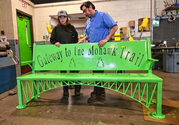Franklin County Technical School senior Laynie Coates, with welding instructor John Passiglia, stands behind her capstone project of a metal bench, with details resembling the French King Bridge, that was mounted on the Erving side of the bridge on Monday.