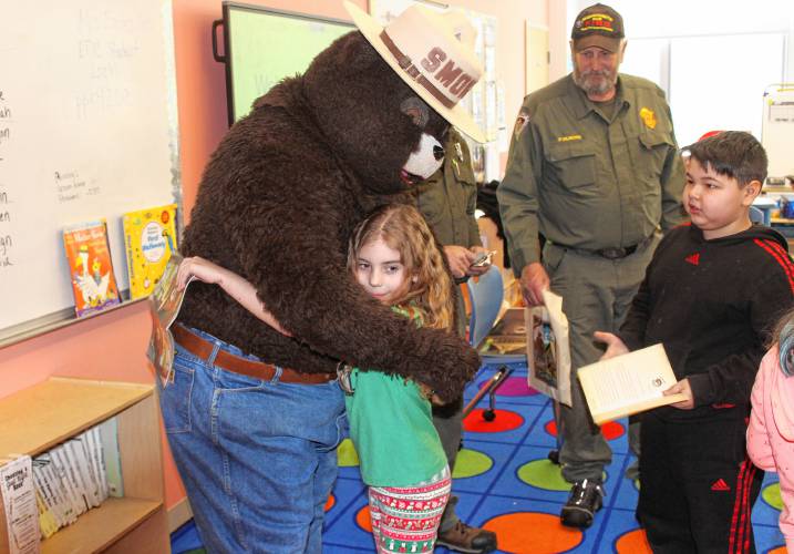Third grader Isabelle Clark gets a hug from Smokey Bear when employees from the state Bureau of Forest Fire Control and Forestry’s location at Erving State Forest visited Fisher Hill Elementary School in Orange on Tuesday as part of Read Across America. District Forest Fire Warden Phil Gilmore stands in the background while classmate Armani Munoz waits his turn in India Eadie’s classroom.