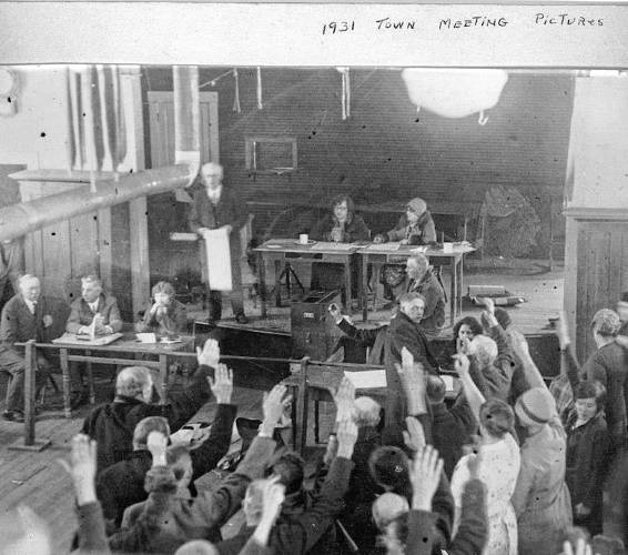 At the 1931 Town Meeting, the stovepipe divides “Bakerites” (seated on the right) from “Ballouites” (seated on the left). Charles Ballou is seated, far left. Town Moderator Ernest Stillman Putnam is standing on stage.